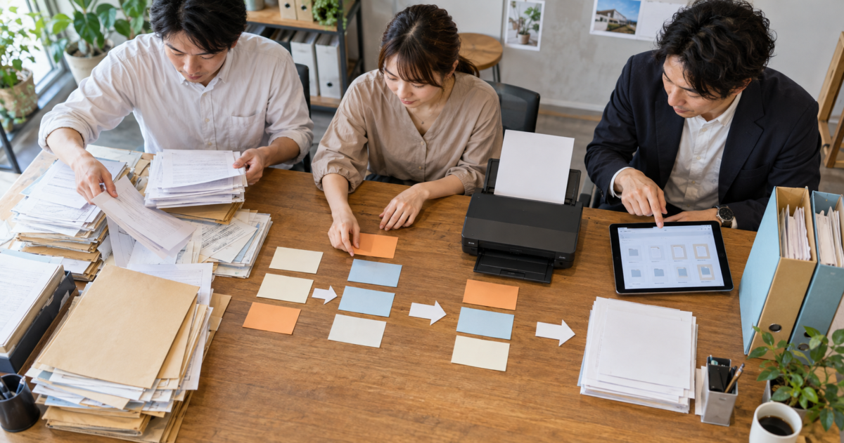 紙書類の山から必要な工程を抜き出しタブレットまでの業務の流れを整理する三人の担当者